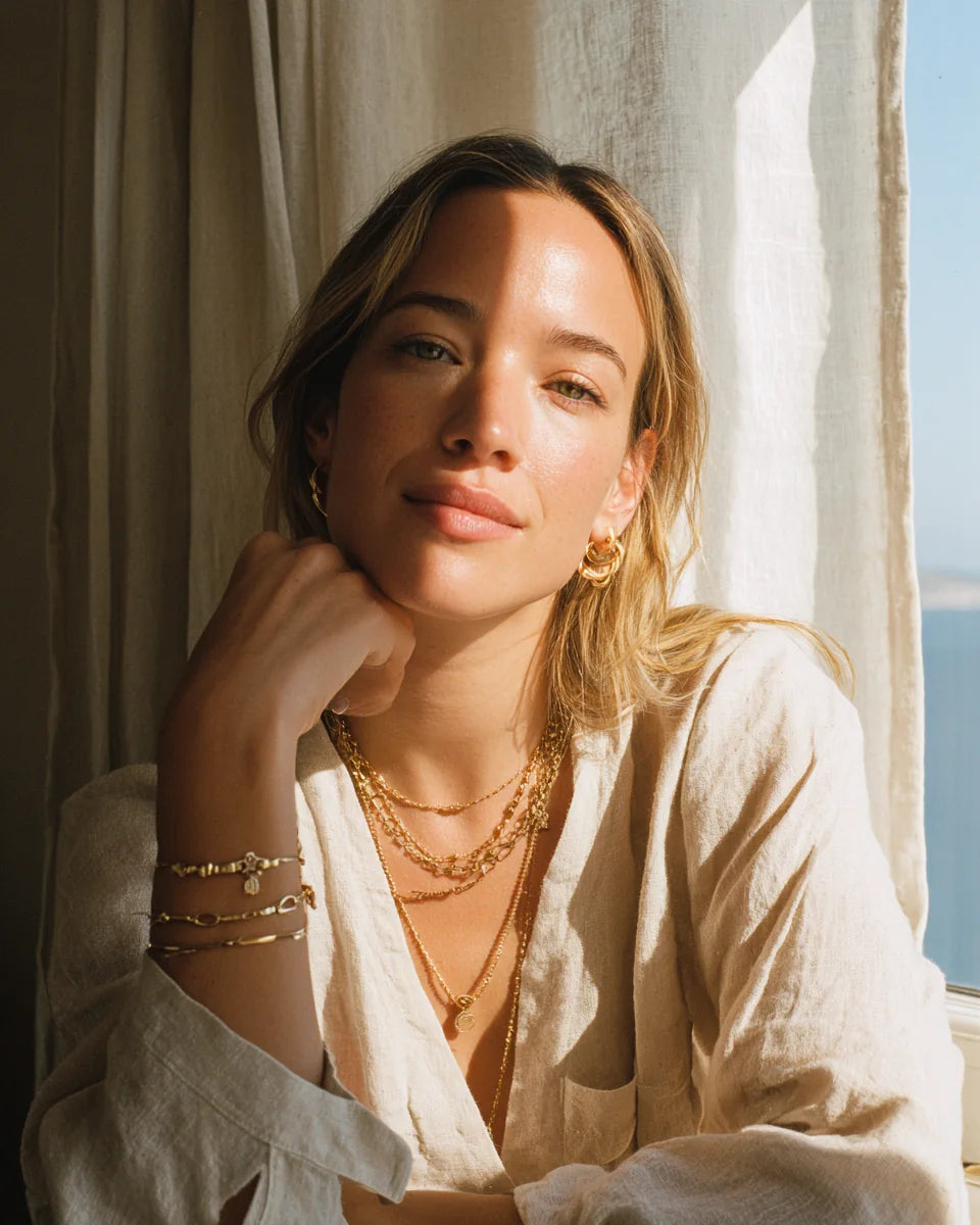 Woman wearing layered gold jewelry beside a sunlit window.