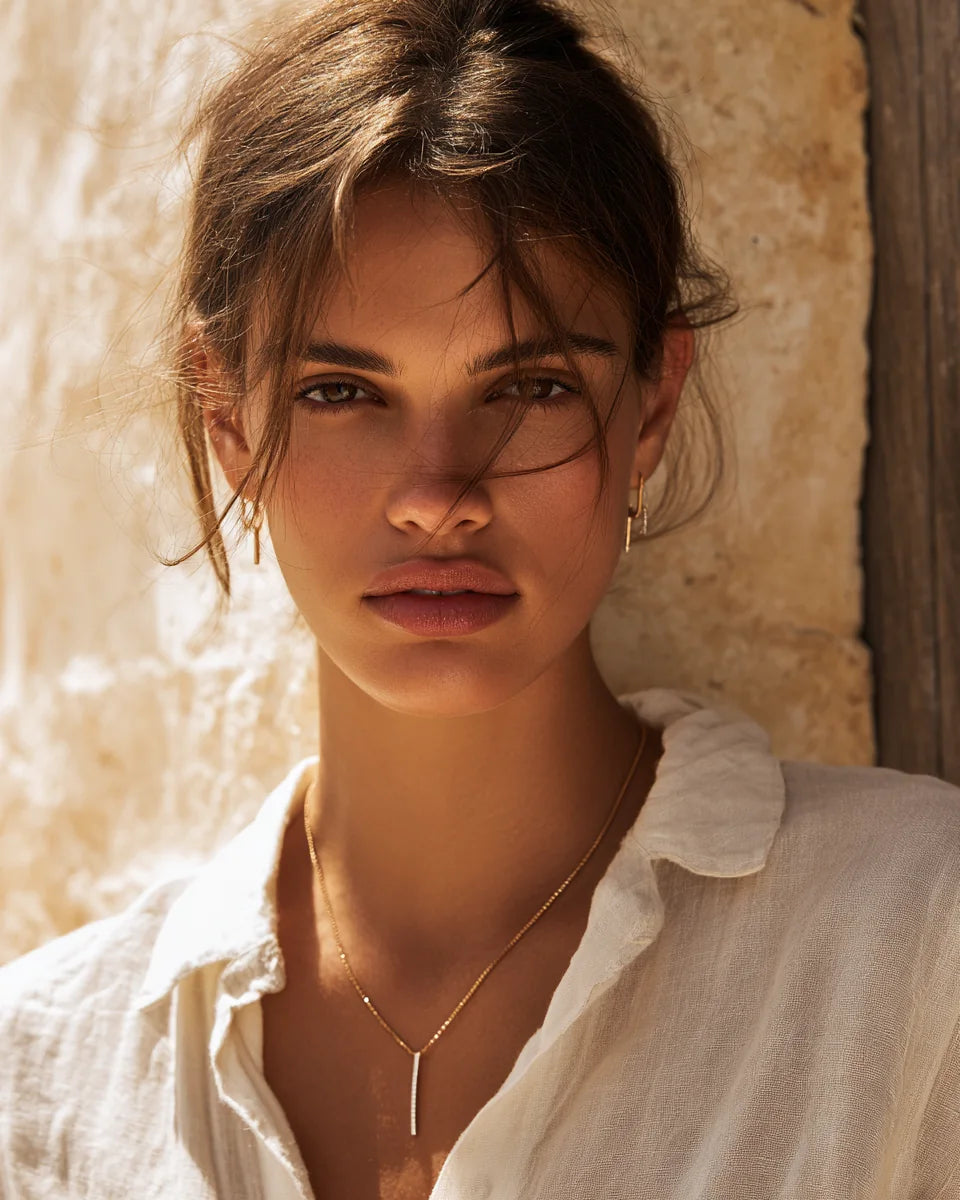 Woman wearing delicate gold jewelry against a warm Mediterranean stone wall.