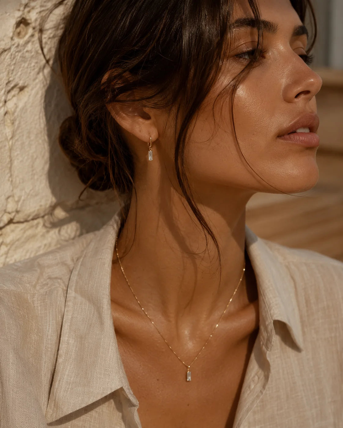 Woman wearing delicate gold jewelry beside a Mediterranean stone wall.