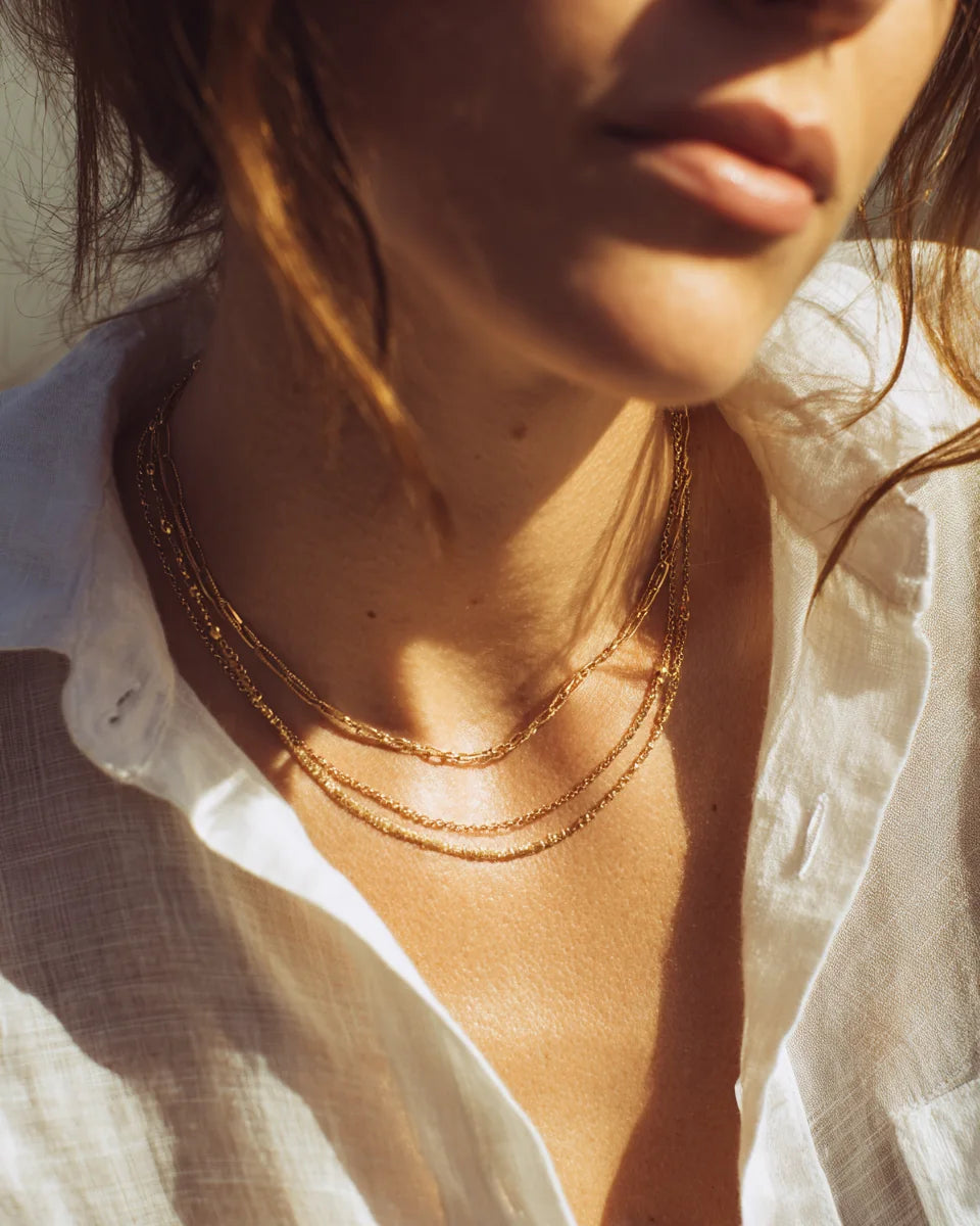 Woman styling layered gold necklaces in warm natural light.